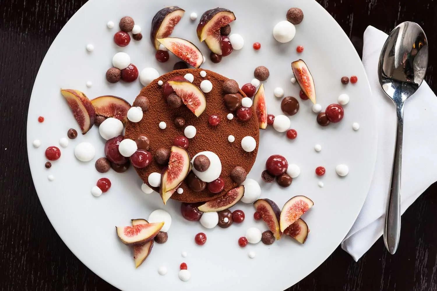 Overhead chocolate mousse cake dusted with cocoa, fresh figs, chocolate pearls, and meringue drops on a white plate