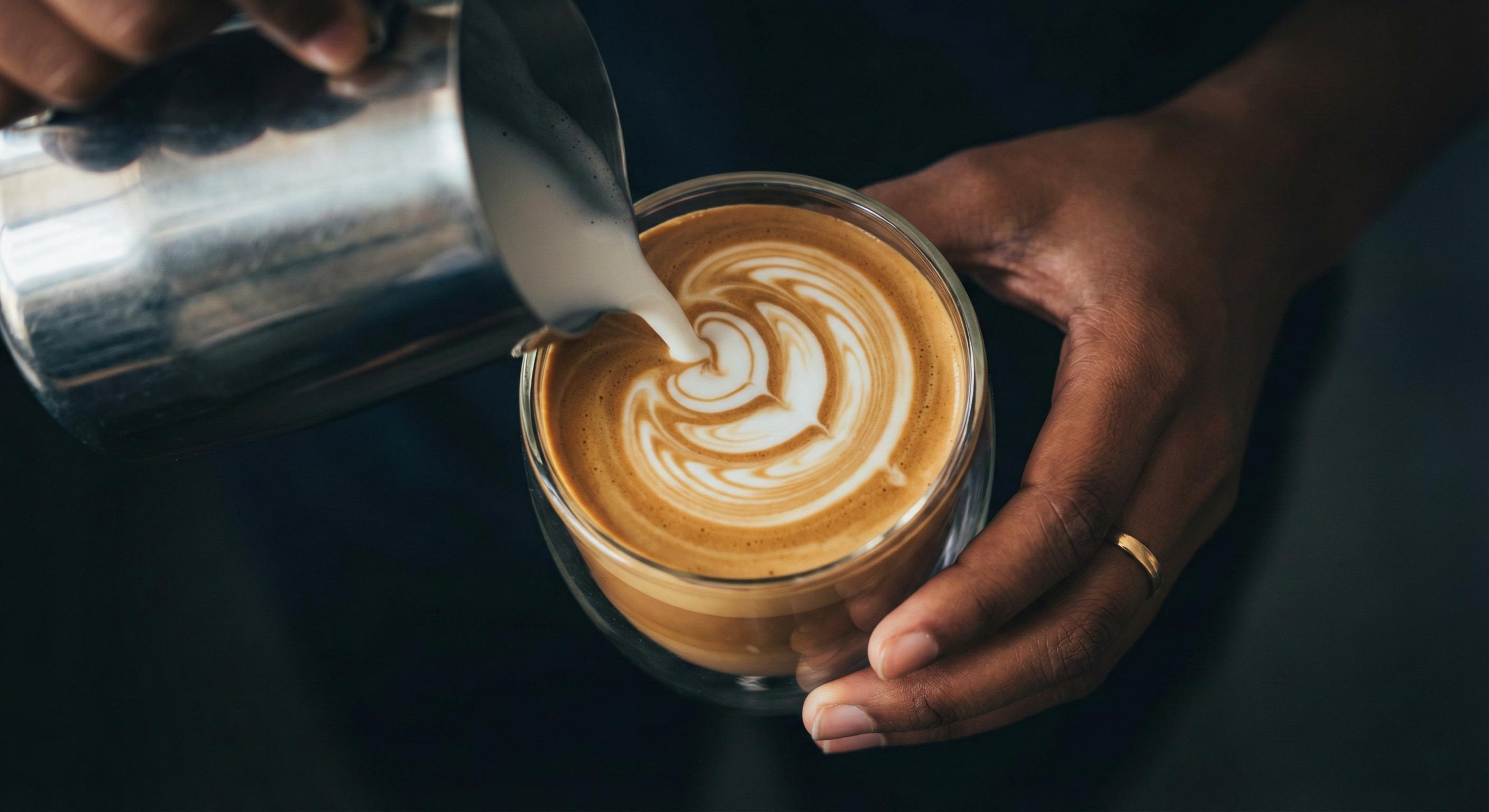 Barista pouring latte art into a glass cup, creating a swirl pattern with steamed milk